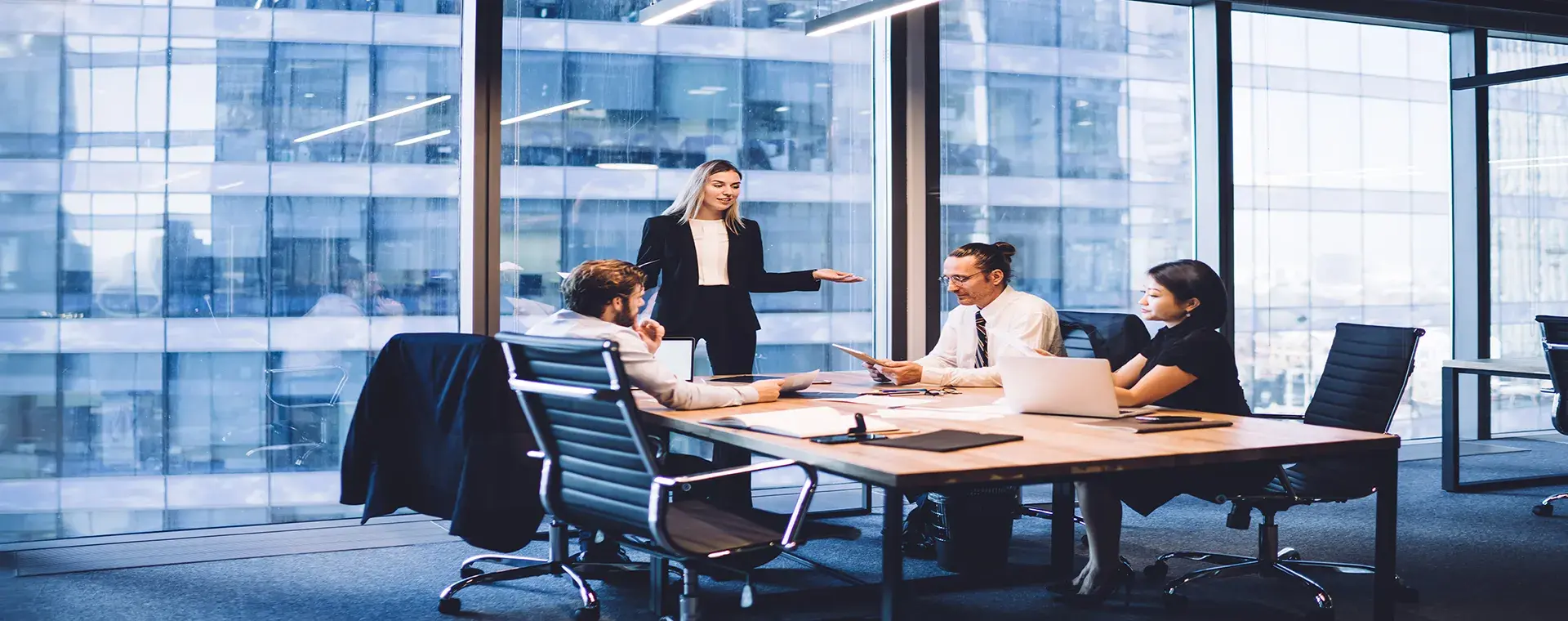Four colleagues meeting around a boardroom table in a glass-walled office tower, one standing and presenting to the others.